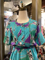 Mannequin wearing a turquoise dress with purple and black abstract pattern in a store setting.